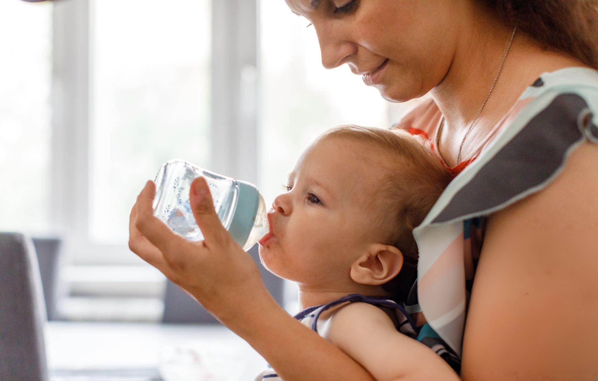 A thirsty boy drinks water from a baby bottle while sitting on his mother's lap. A young mother exercising in her home on an exercise bike - Postpartum Exercise